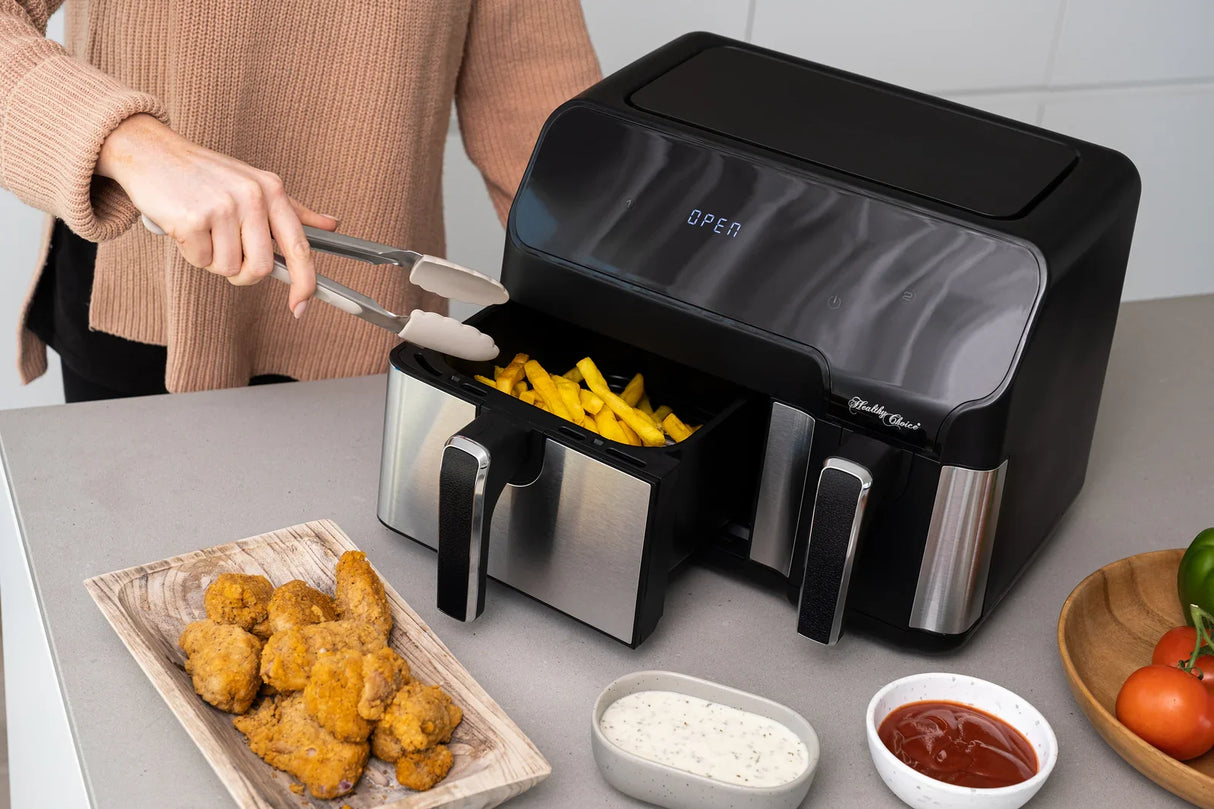 Person using stainless steel tongs to take fries from a black and silver air fryer with fried chicken and dipping sauces on the counter