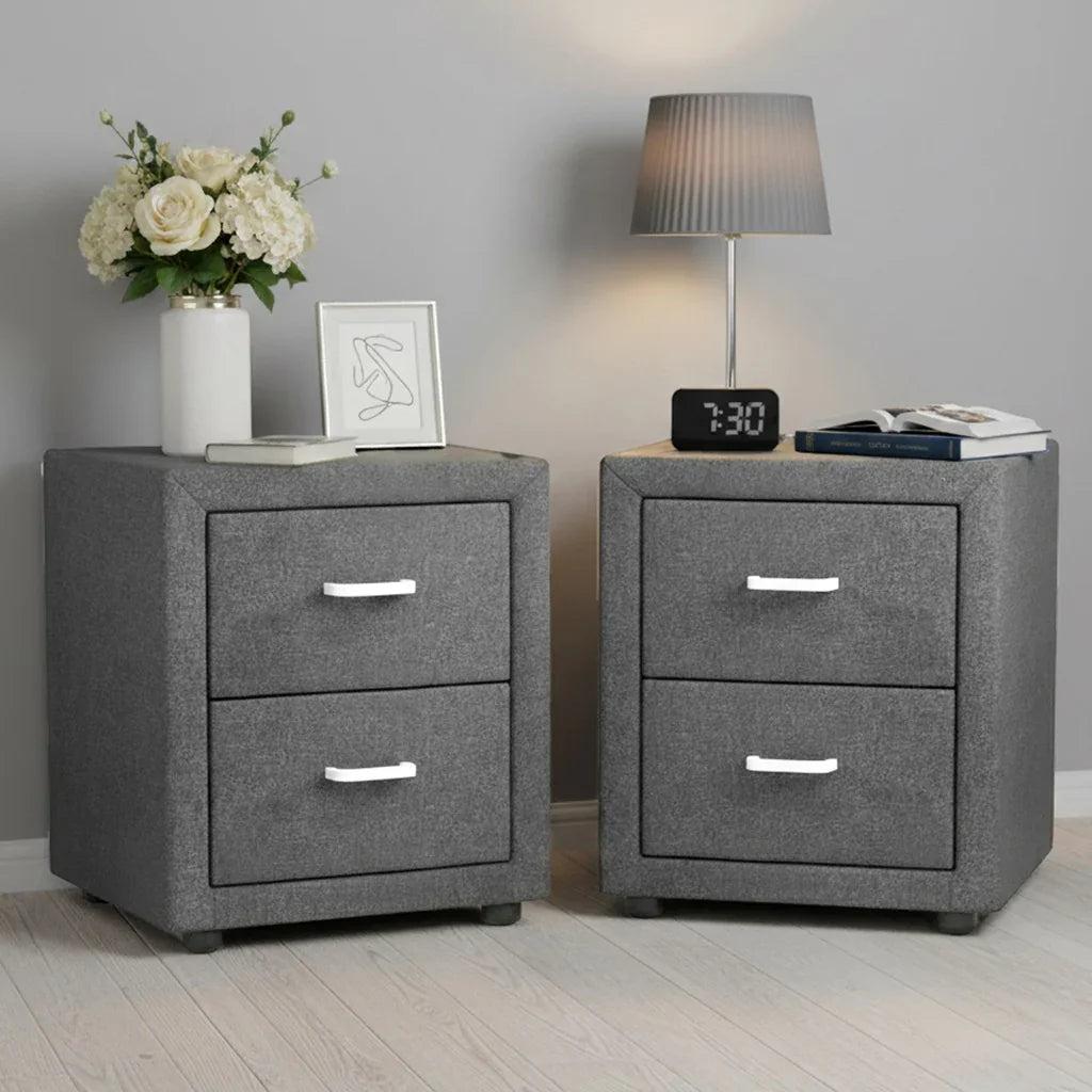 Pair of modern grey fabric bedside tables with drawers, lamp, flowers, books, and alarm clock.