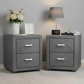 Pair of modern grey fabric bedside tables with drawers, lamp, flowers, books, and alarm clock.