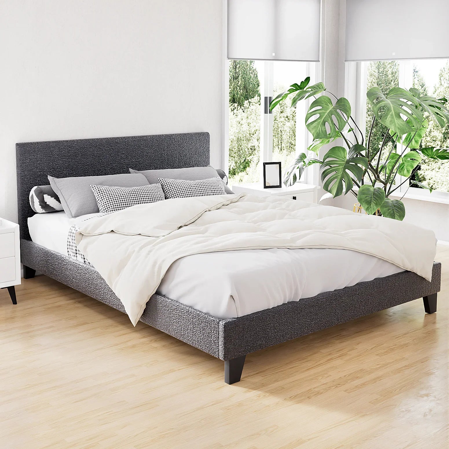 Modern grey upholstered platform bed with white bedding, pillows, and large leafy green plant