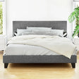 Modern gray upholstered bed with white bedding, decorative pillows, wooden floor, and green plants in a bright room