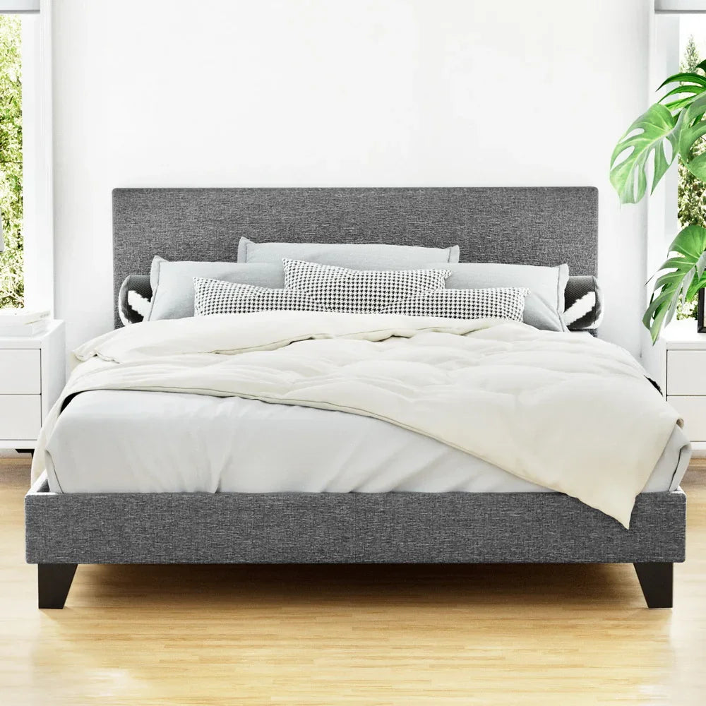 Modern gray upholstered bed with white bedding, decorative pillows, wooden floor, and green plants in a bright room