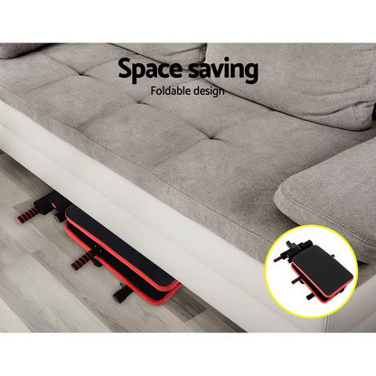 Foldable sit-up bench stored under gray sofa, space-saving home gym equipment.