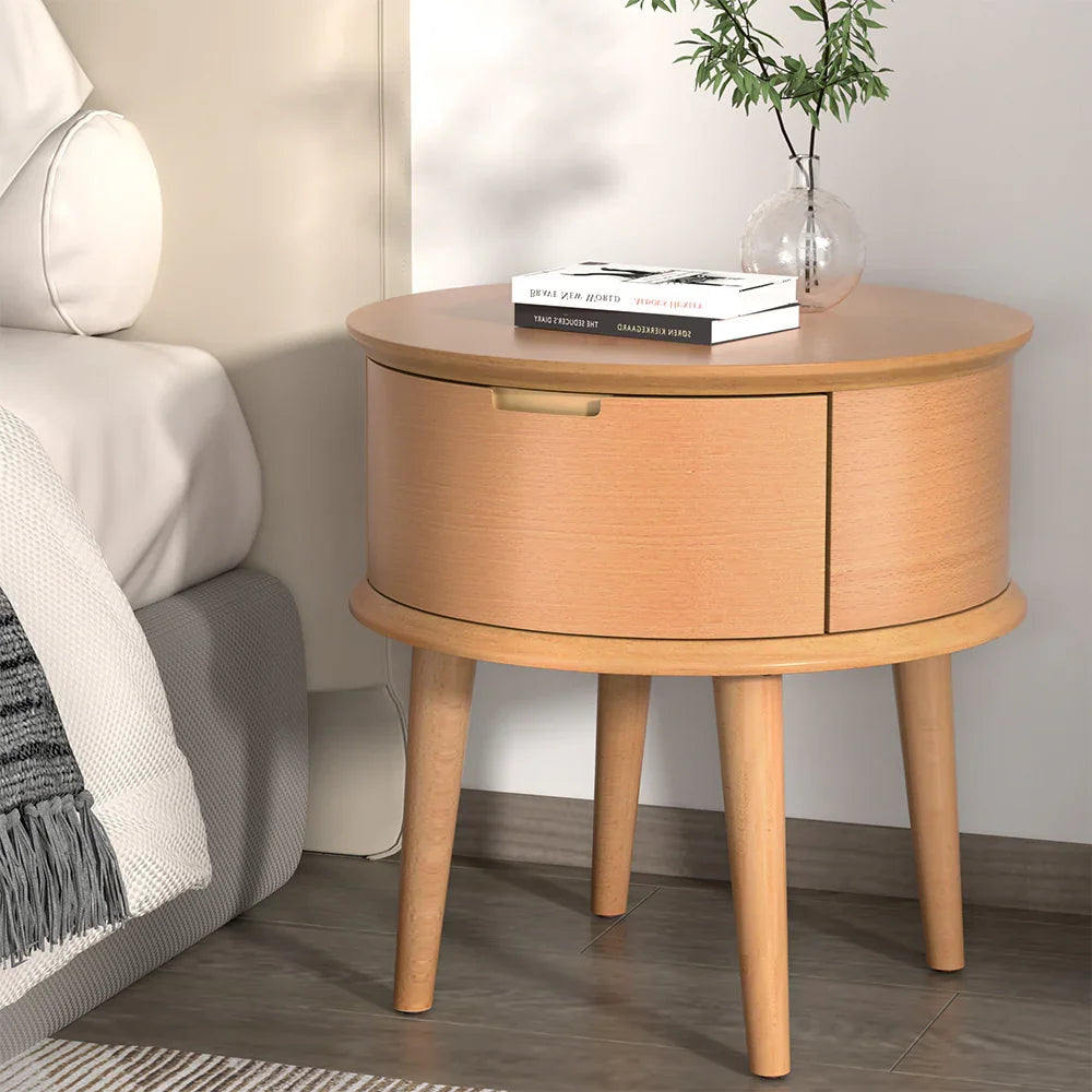 Round wooden bedside table with drawer, books, and small vase next to bed