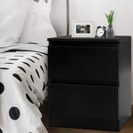 Black bedside table with two drawers, next to a polka dot bed, with clock, vase, and photo frame on top