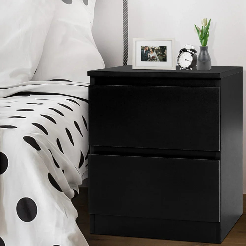 Black bedside table with two drawers, next to a polka dot bed, with clock, vase, and photo frame on top