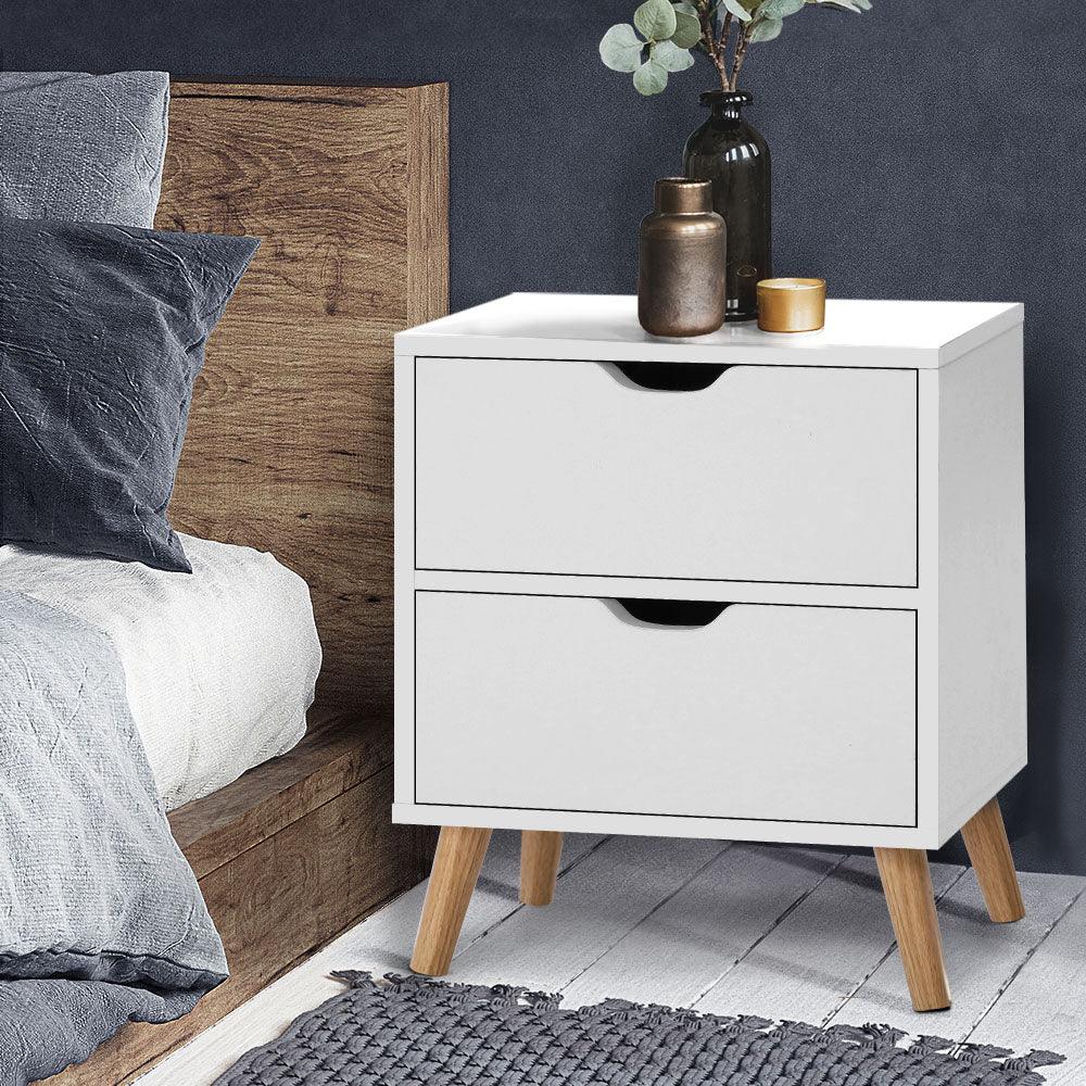 Modern white two-drawer bedside table with wooden legs next to a bed in a cozy bedroom