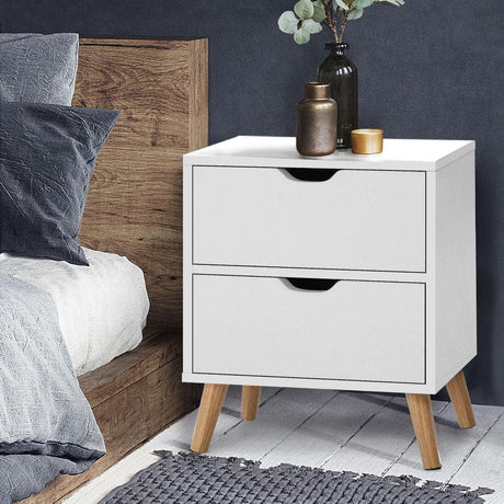 Modern white two-drawer bedside table with wooden legs next to a bed in a cozy bedroom