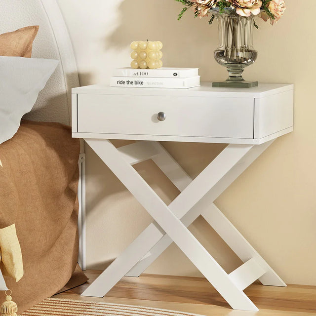 Modern white nightstand with crossed legs, decorative candle, books, and flower vase
