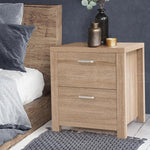 Modern light wood bedside table with two drawers, metal handles, and decorative vases.