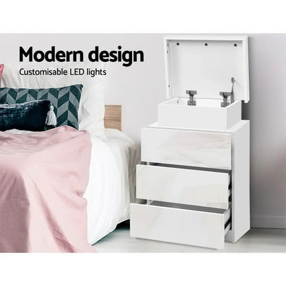 Modern white bedside table with LED lights and open drawers beside a bed with pink bedding.