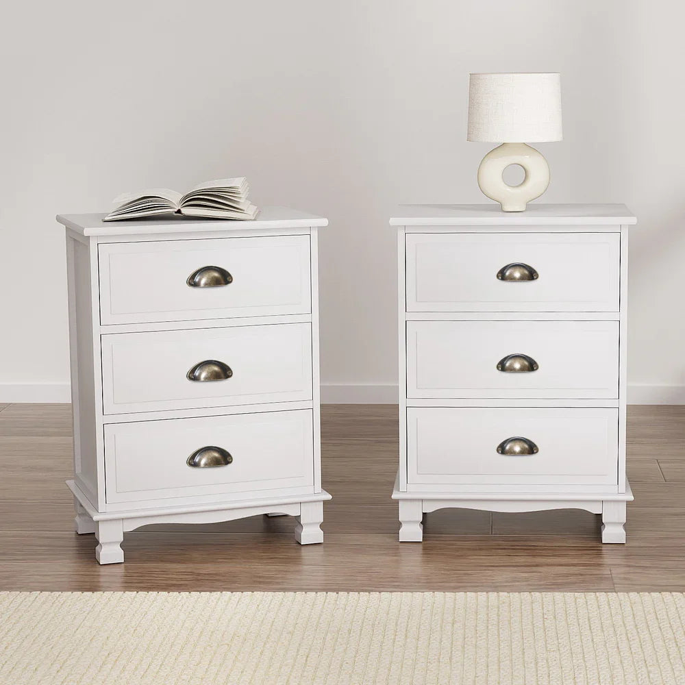 Pair of white three-drawer nightstands with brass handles, lamp, and book on top