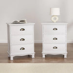 Pair of white three-drawer nightstands with brass handles, lamp, and book on top