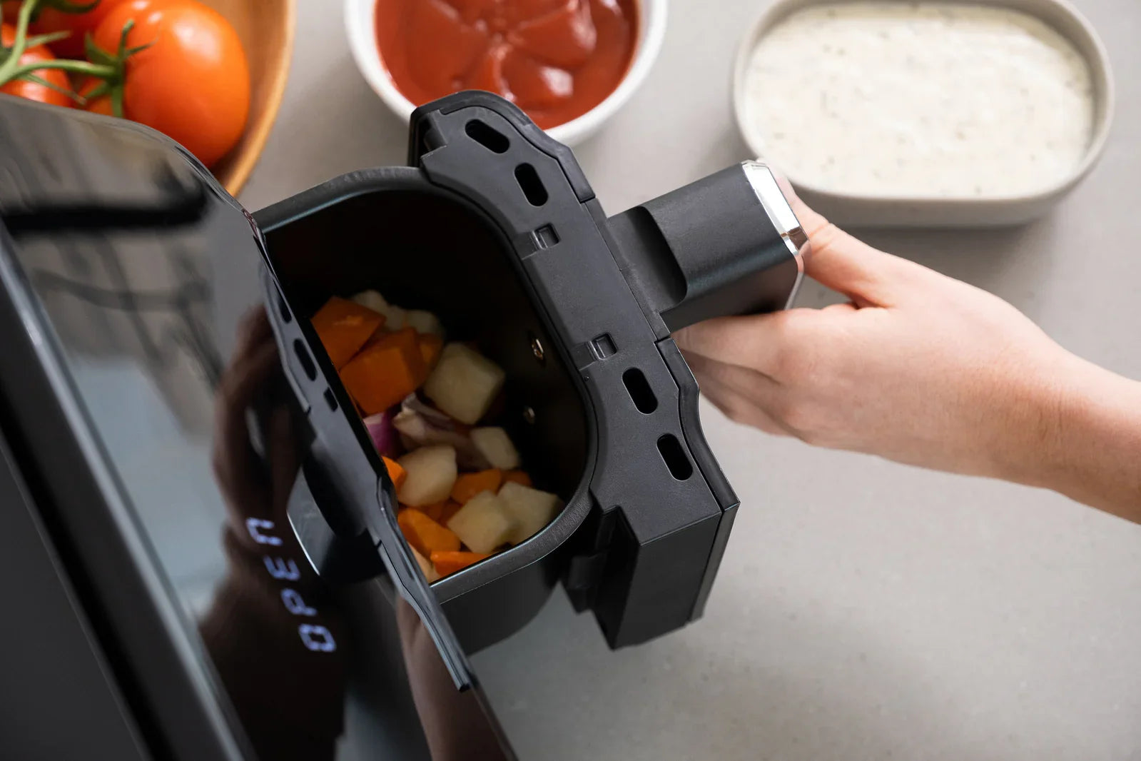 Hand removing air fryer basket filled with chopped vegetables on kitchen counter with sauces and tomatoes nearby