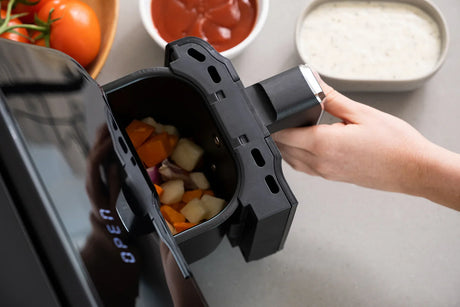 Hand removing air fryer basket filled with chopped vegetables on kitchen counter with sauces and tomatoes nearby