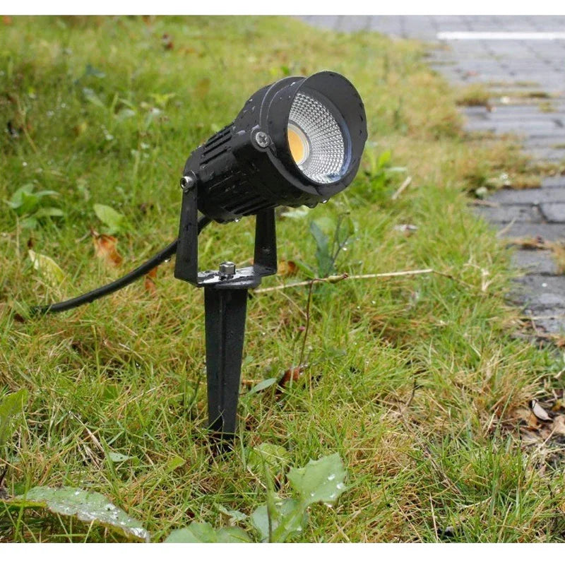 Black outdoor garden spotlight fixture installed in grassy area near a paved walkway
