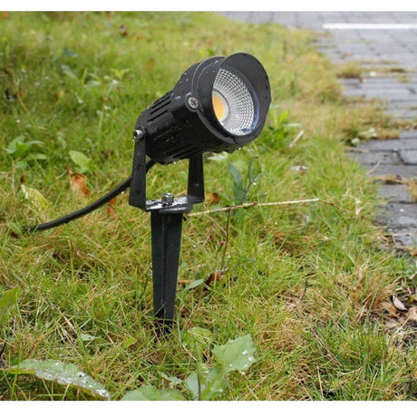 Black outdoor garden spotlight fixture installed in grassy area near a paved walkway