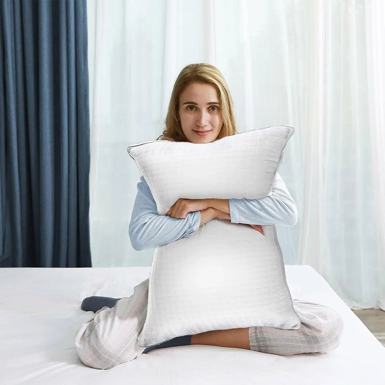 Smiling woman in pajamas sitting on bed hugging large white textured pillow in bright bedroom
