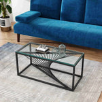 Modern glass coffee table with geometric black metal base in front of a blue velvet sofa