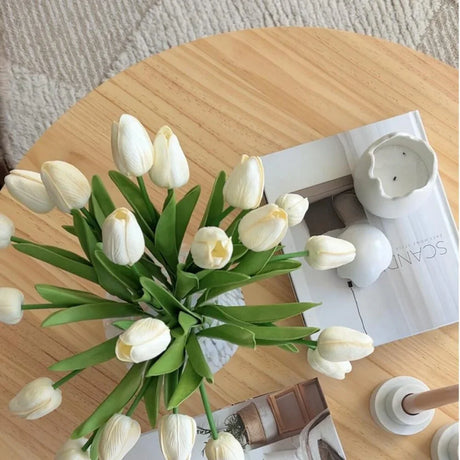 White tulips in vase on wooden table with candle, magazine, and modern decor