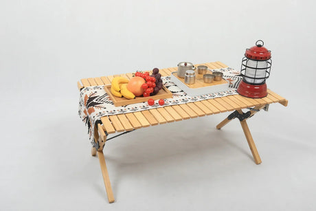 Portable wooden folding camping table with fruit tray, tea set, and red lantern on patterned cloth