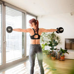 Woman in black sports bra and gray leggings lifting dumbbells in bright living room with plants and TV