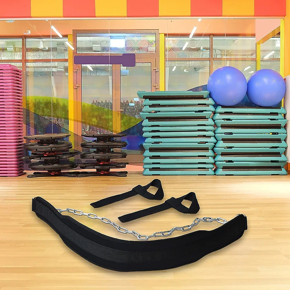 Gym equipment including black dip belt with chain, ankle straps on wooden floor, stacked aerobic steps, and blue exercise balls