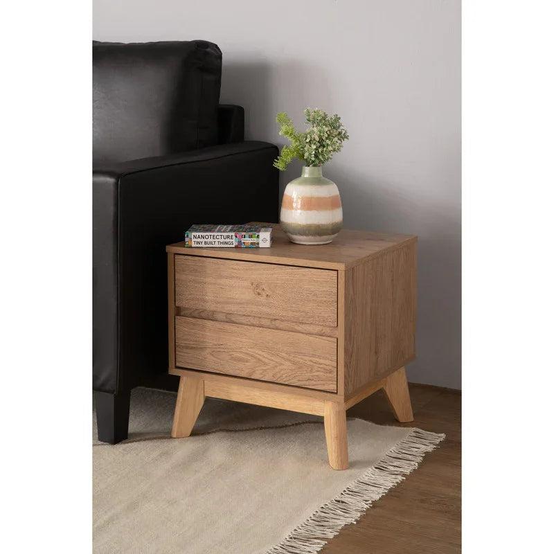 Modern wooden nightstand with two drawers, books, and a vase with greenery beside black sofa