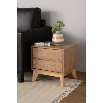Modern wooden nightstand with two drawers, books, and a vase with greenery beside black sofa