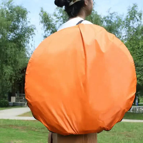 Person outdoors carrying large round bright orange backpack cover with greenery and path in background