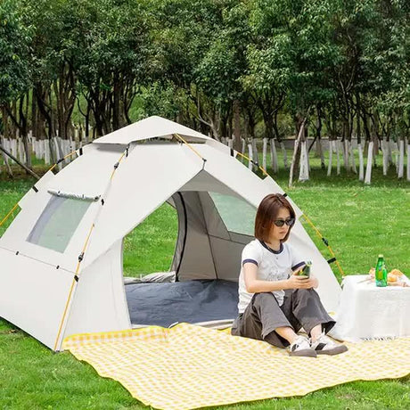 Woman sitting on a yellow checkered picnic blanket outside a white camping tent in a green park with trees