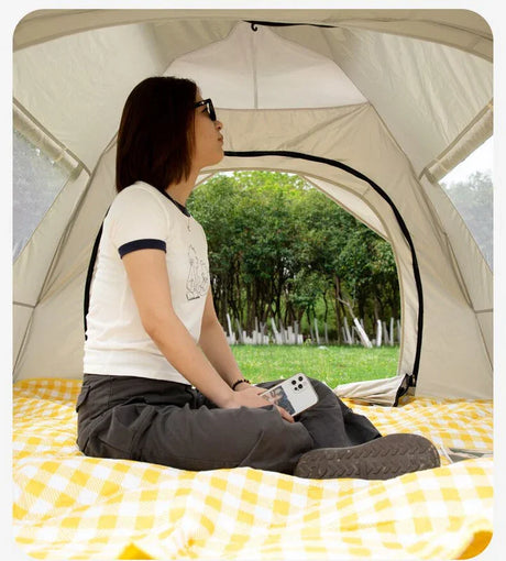 Woman sitting inside beige tent on yellow checkered blanket holding smartphone, green trees visible outside