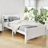 White wooden bed with gray bedding and decorative pillows in bright room with large windows and green plant